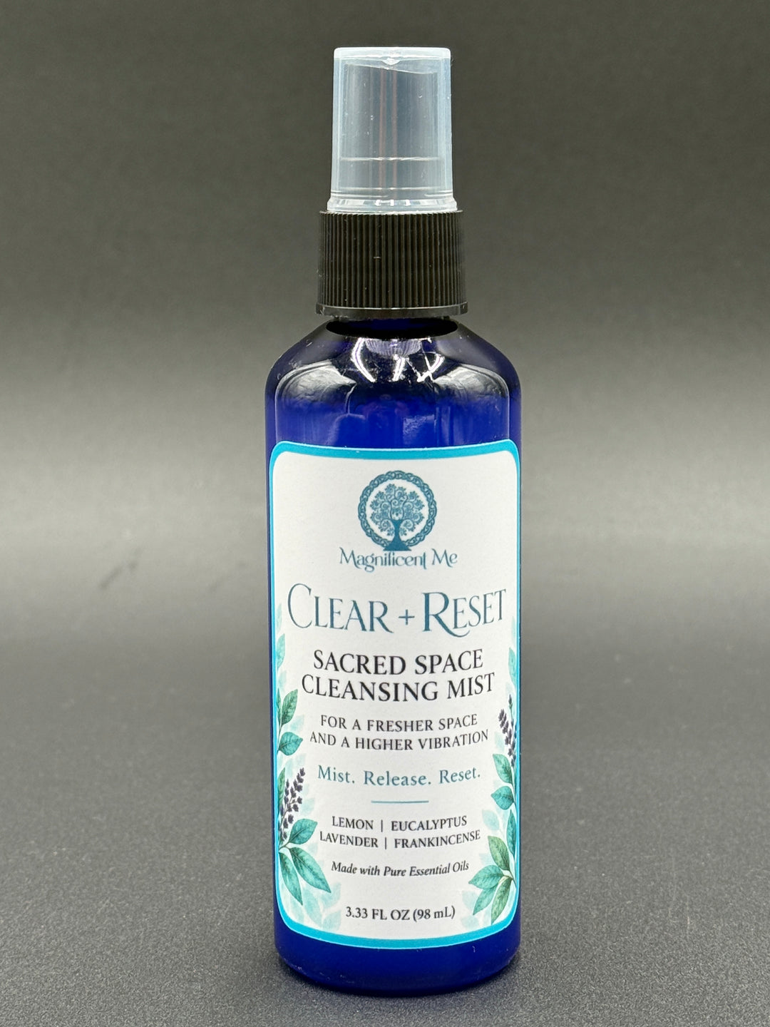 Magnificent Me Clear + Reset essential oil room spray in a cobalt-blue bottle with botanical label, featuring lemon, eucalyptus, lavender and frankincense.