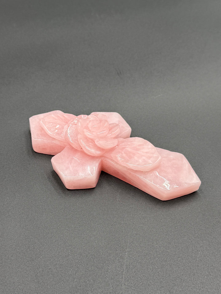Detailed view of a natural pink rose quartz cross showing the smooth polished crystal surface, subtle white inclusions, and sculpted rose petals.
