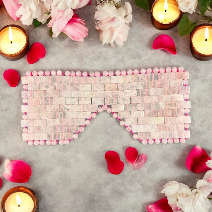Natural rose quartz crystal eye mask displayed with candles and rose petals in a luxury spa-inspired self-care setting for relaxation and wellness.